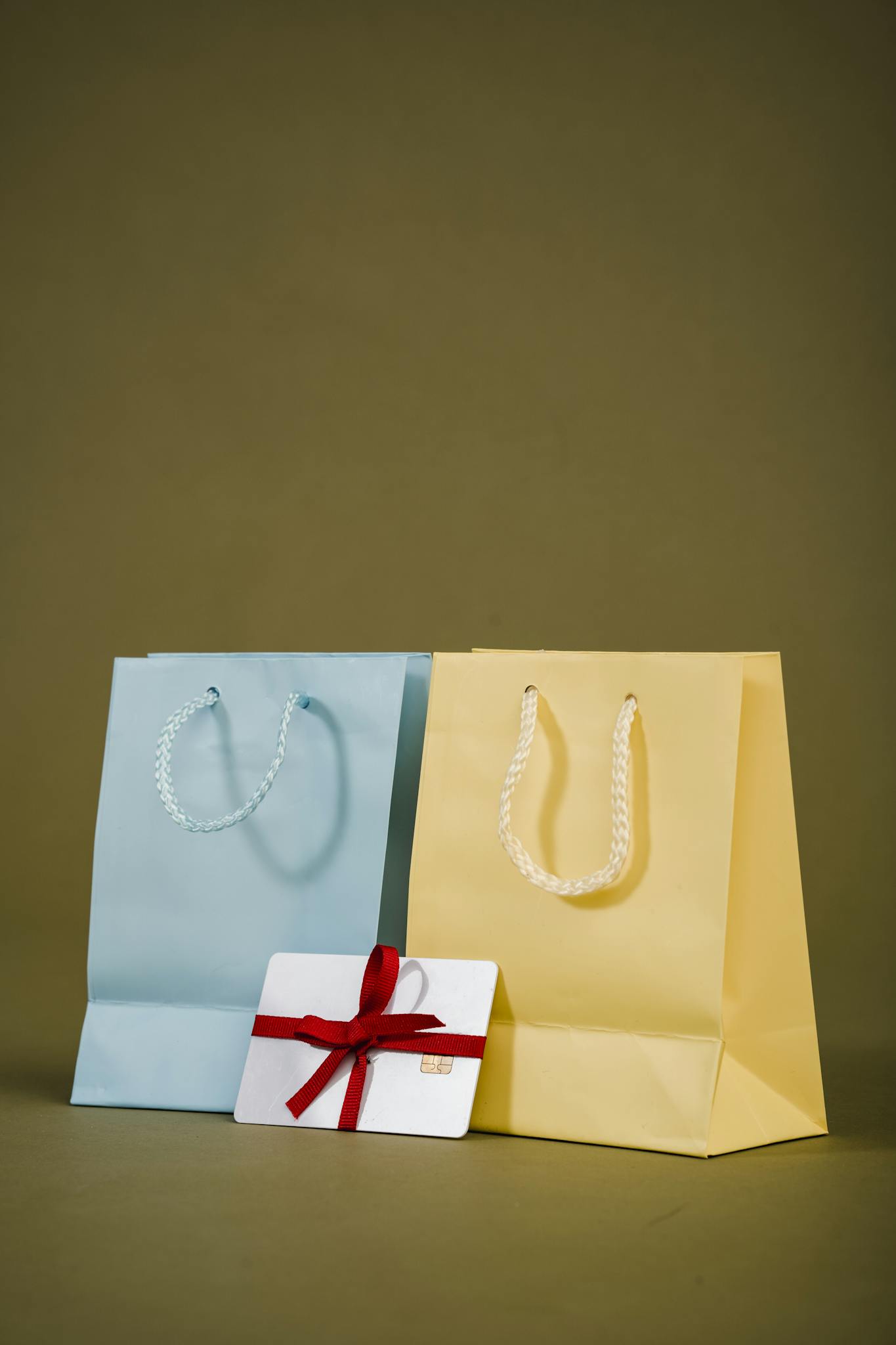 Pastel blue and yellow shopping bags with a red-ribboned gift card on an olive background, ideal for retail themes.