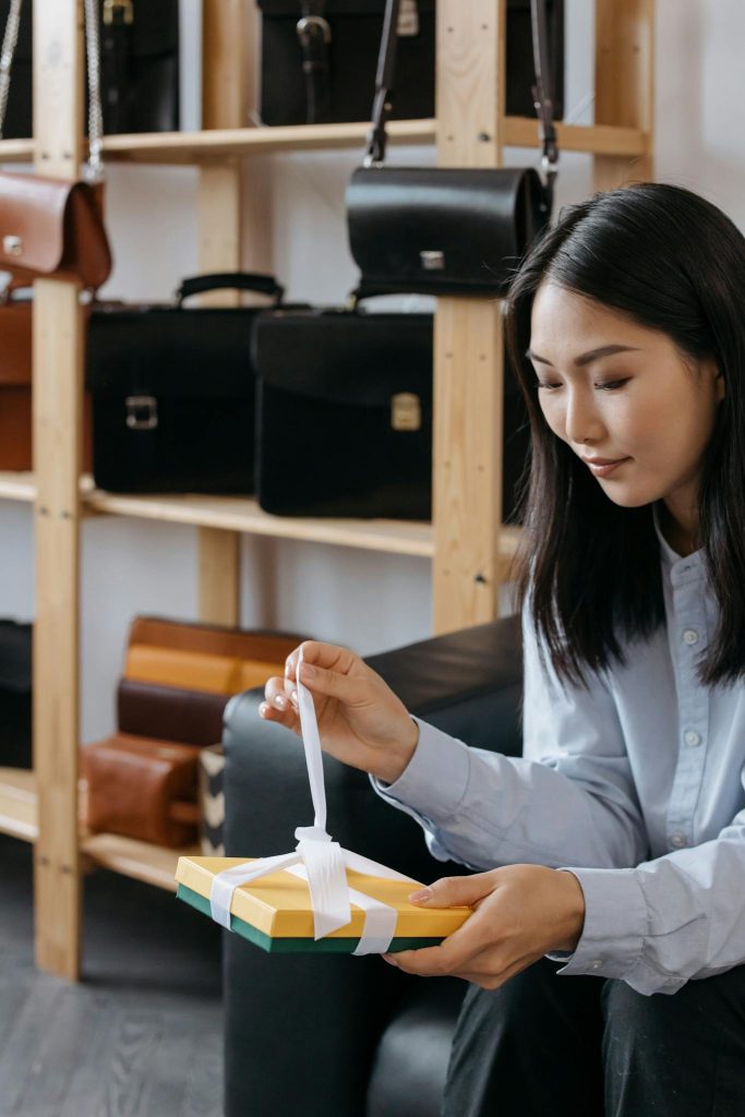 Woman sitting indoors unwrapping a gift box surrounded by leather bags.